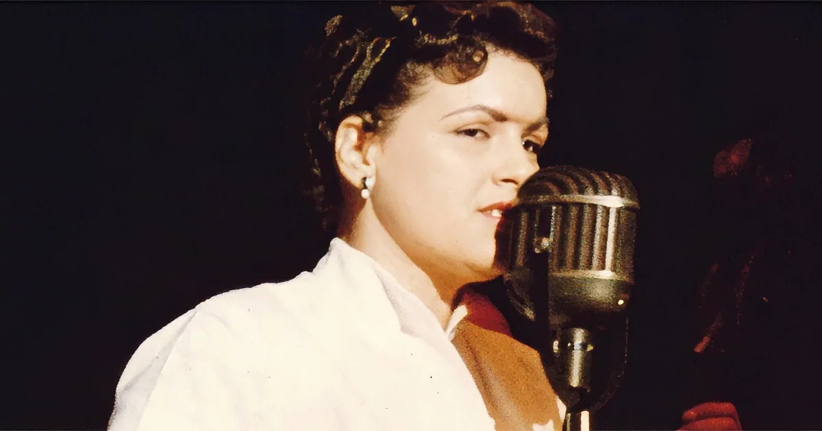 A woman in a white shirt sings into a vintage microphone, her hair styled in a 1950s fashion.