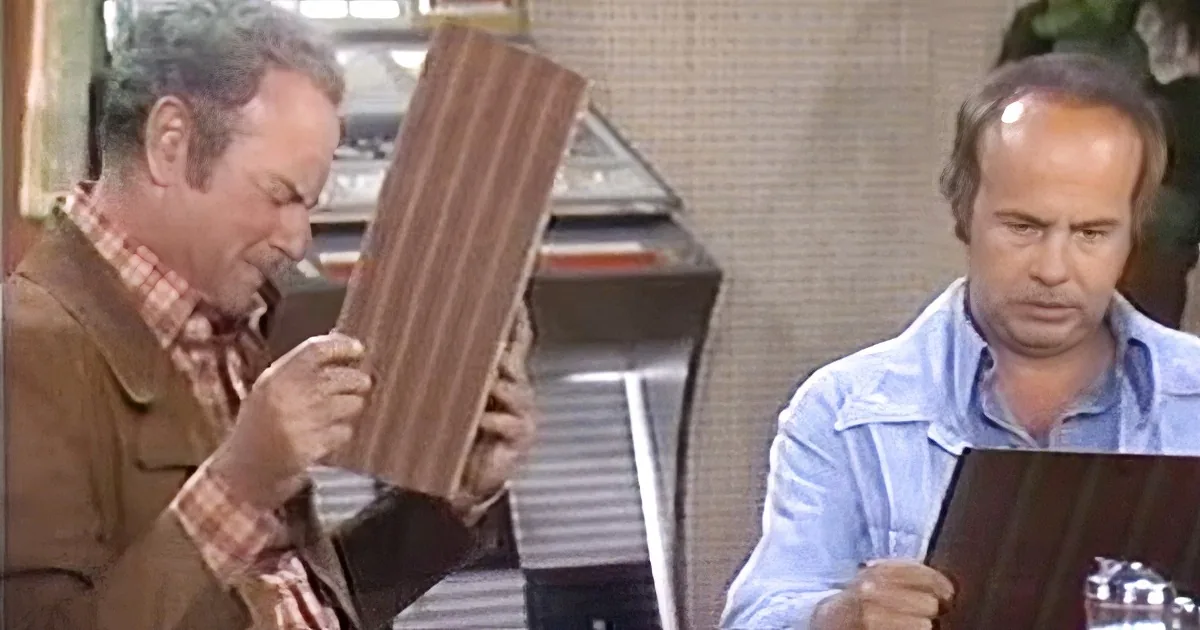 A man in a brown jacket holds a wooden object while another man in a blue shirt looks on.