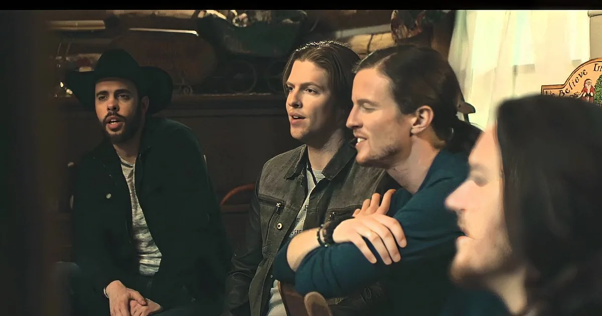 Three men in a dimly lit room, one wearing a cowboy hat, engaged in conversation.