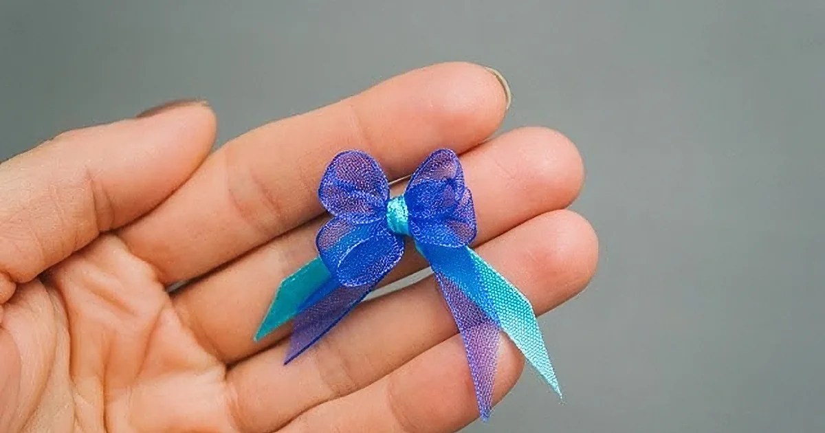 A hand holds a small, intricately tied blue ribbon bow against a plain background.
