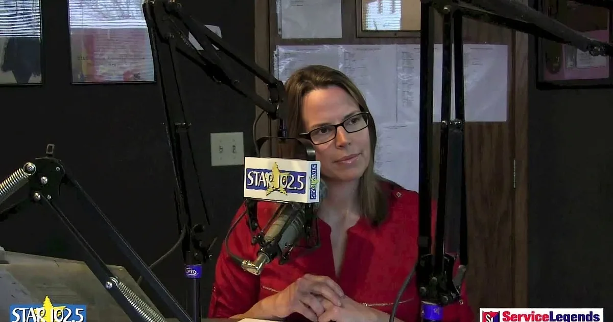 A woman in a red shirt sits at a radio station desk with a microphone, surrounded by equipment and promotional signs.