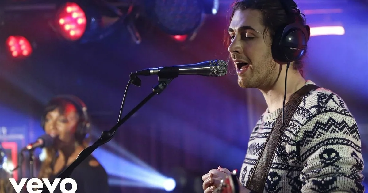 A man wearing headphones and a patterned sweater sings into a microphone while holding a guitar.