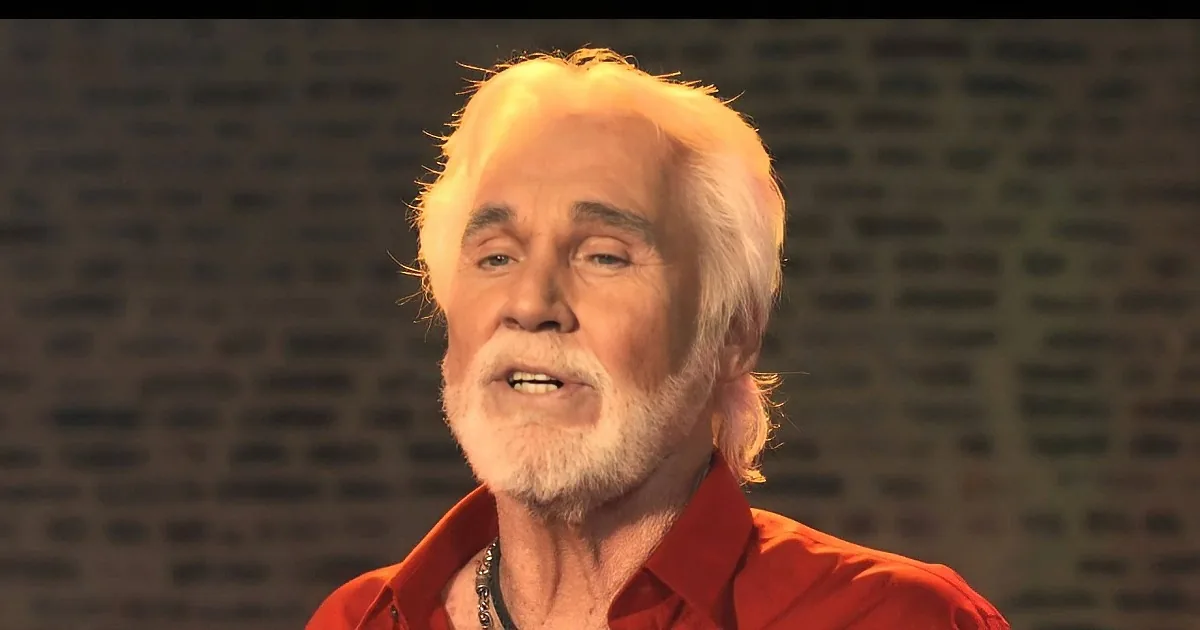 An older man with white hair and a beard, wearing a red shirt, is seen against a brick wall background.