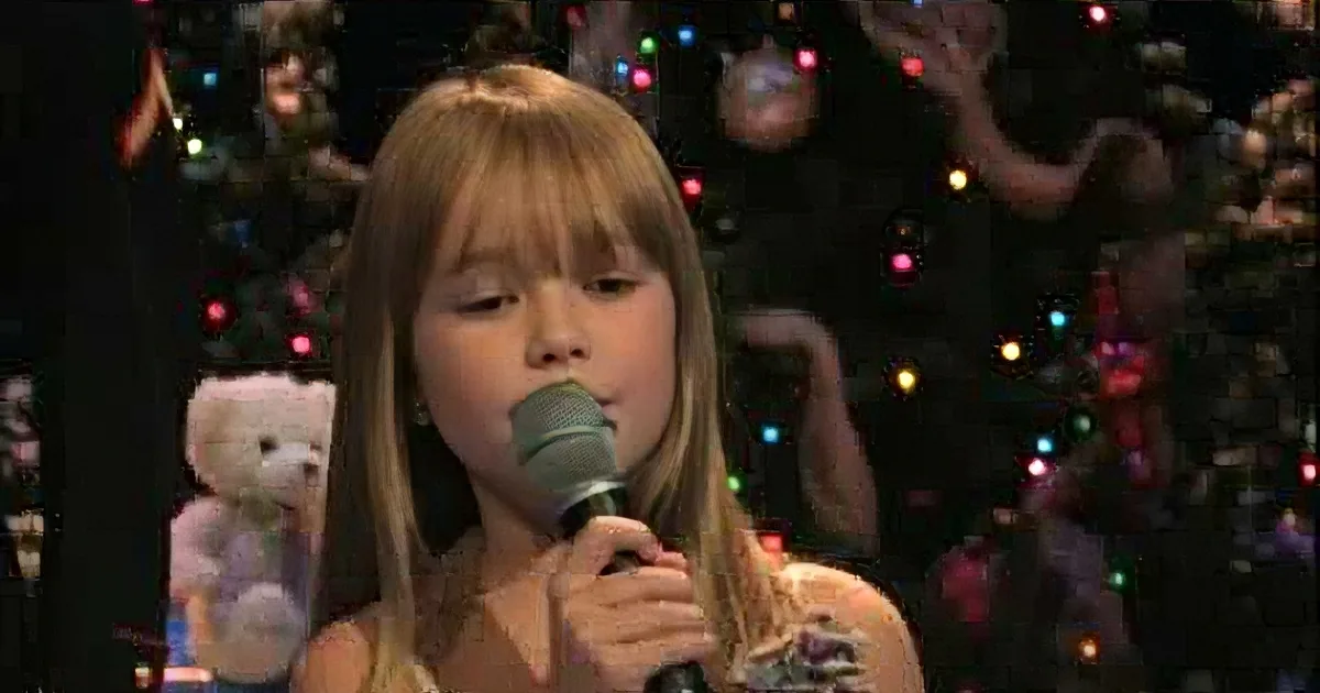 A young girl with long hair sings into a microphone, surrounded by Christmas decorations and teddy bears.