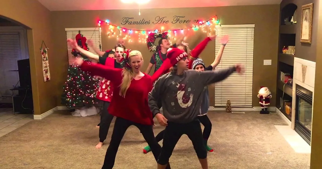 A group of people in festive attire dances joyfully in a decorated living room with Christmas lights and a fireplace.