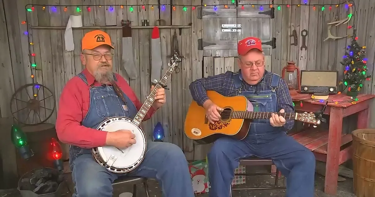 Two men in overalls play banjo and guitar in a rustic setting with Christmas lights.