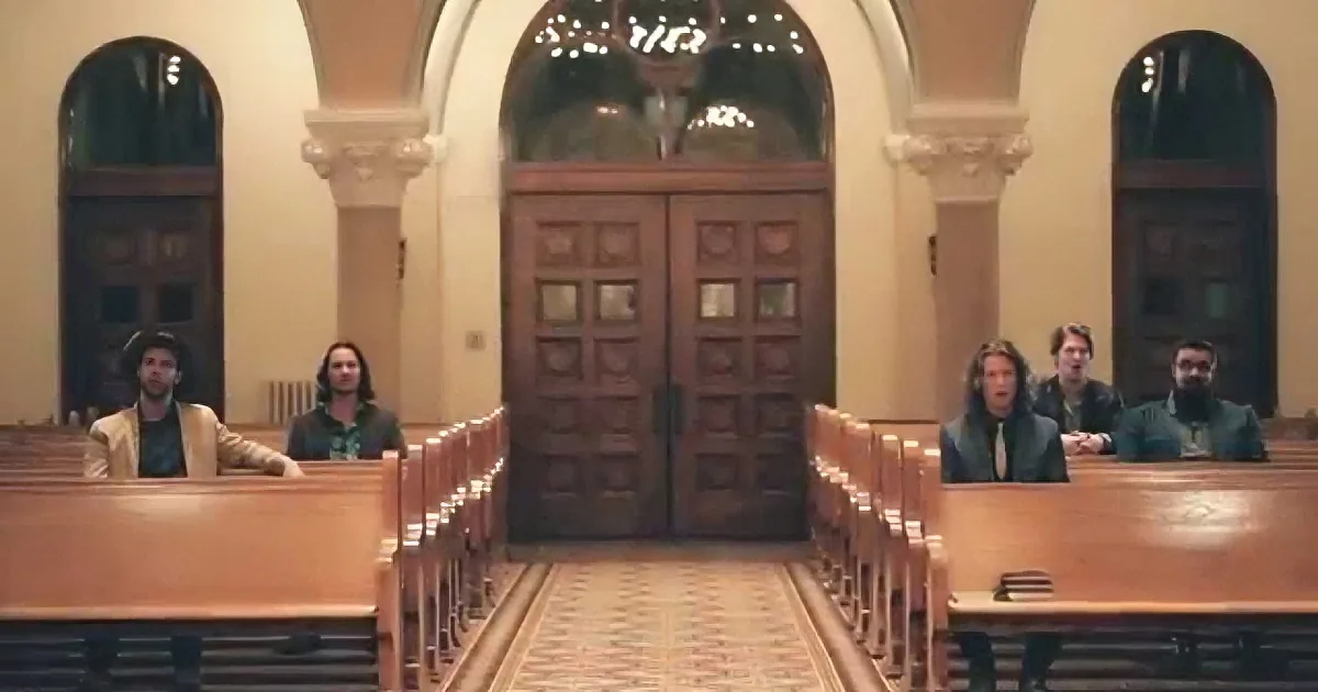 People sit in pews in a church with ornate architecture and arched windows.