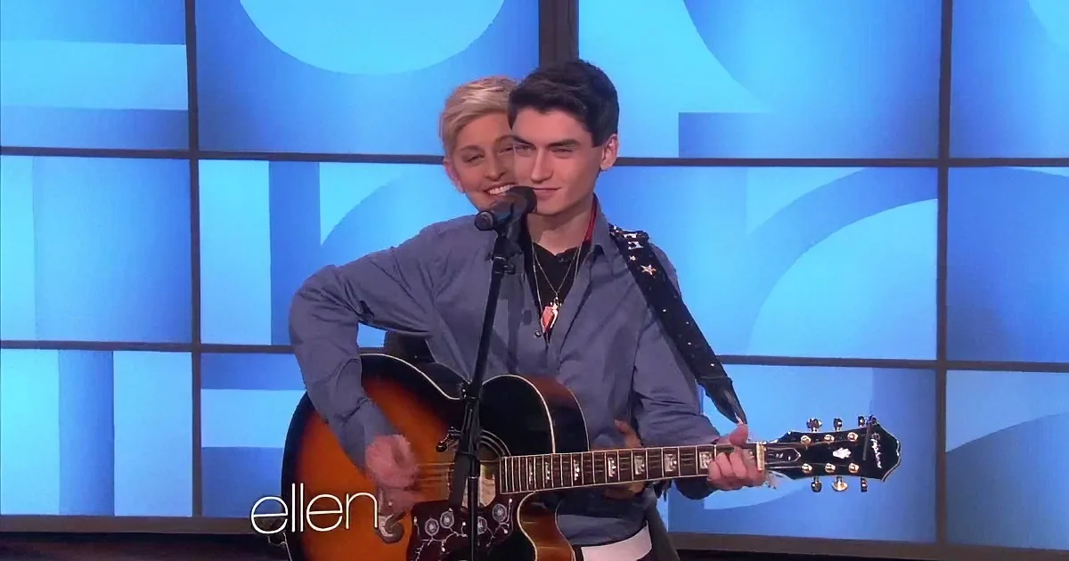 A man plays guitar on stage with a microphone, smiling, in front of a blue background.