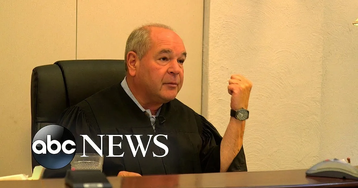 A man in a judge's robe sits at a desk, gesturing with his right hand, in a courtroom setting.