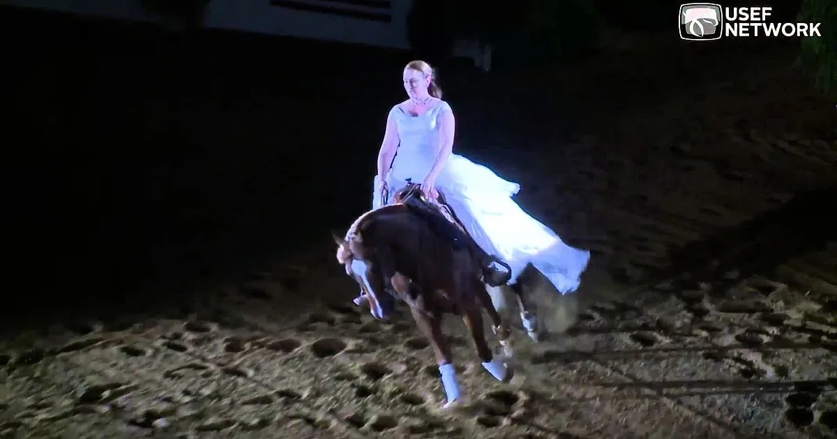 A woman in a white dress rides a horse on a sandy arena at night.