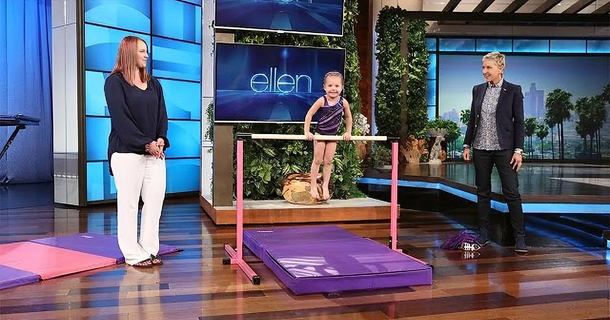 A young girl stands on a balance beam on a TV show set, with two adults nearby.