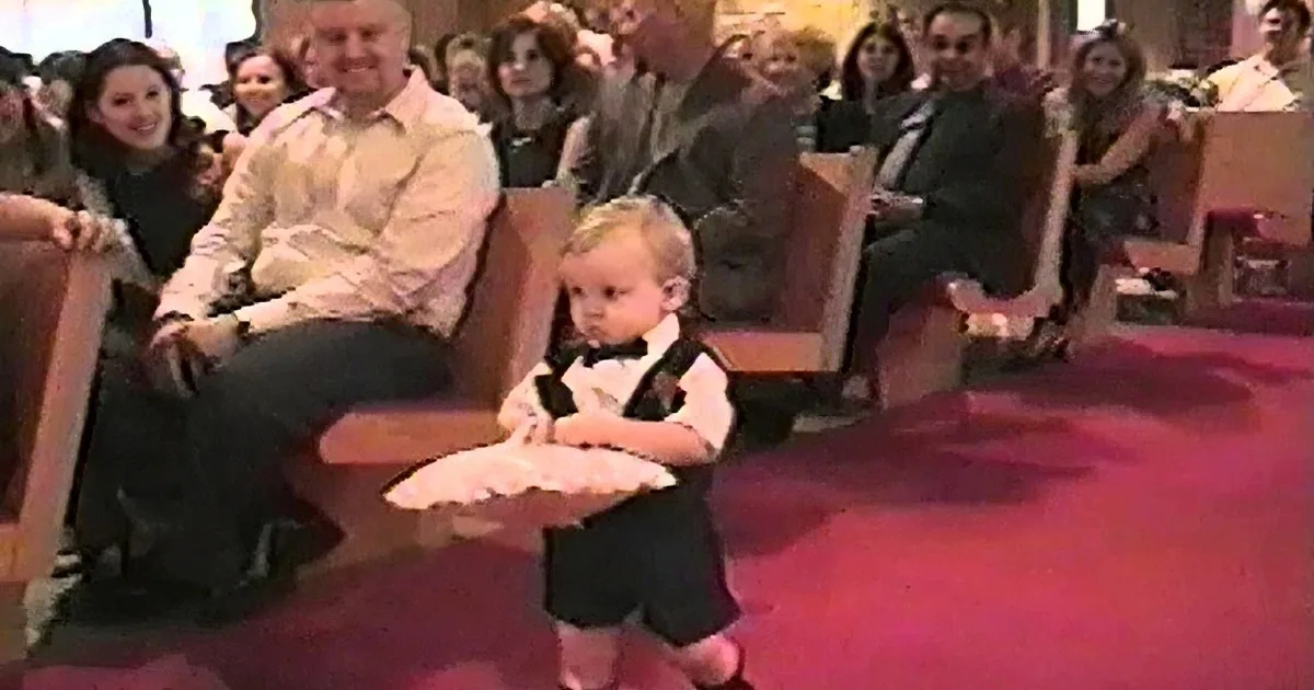 A toddler in a tuxedo sits on a red carpet, surrounded by seated adults in a formal setting.