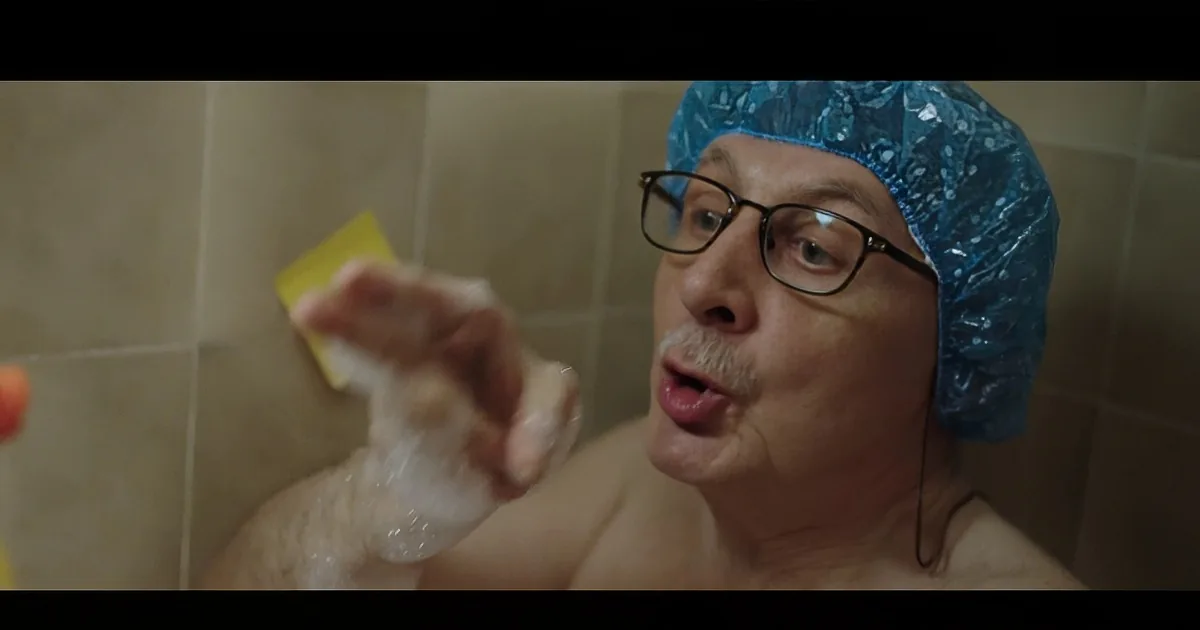 A man wearing a shower cap and glasses is shaving his face in a bathroom.