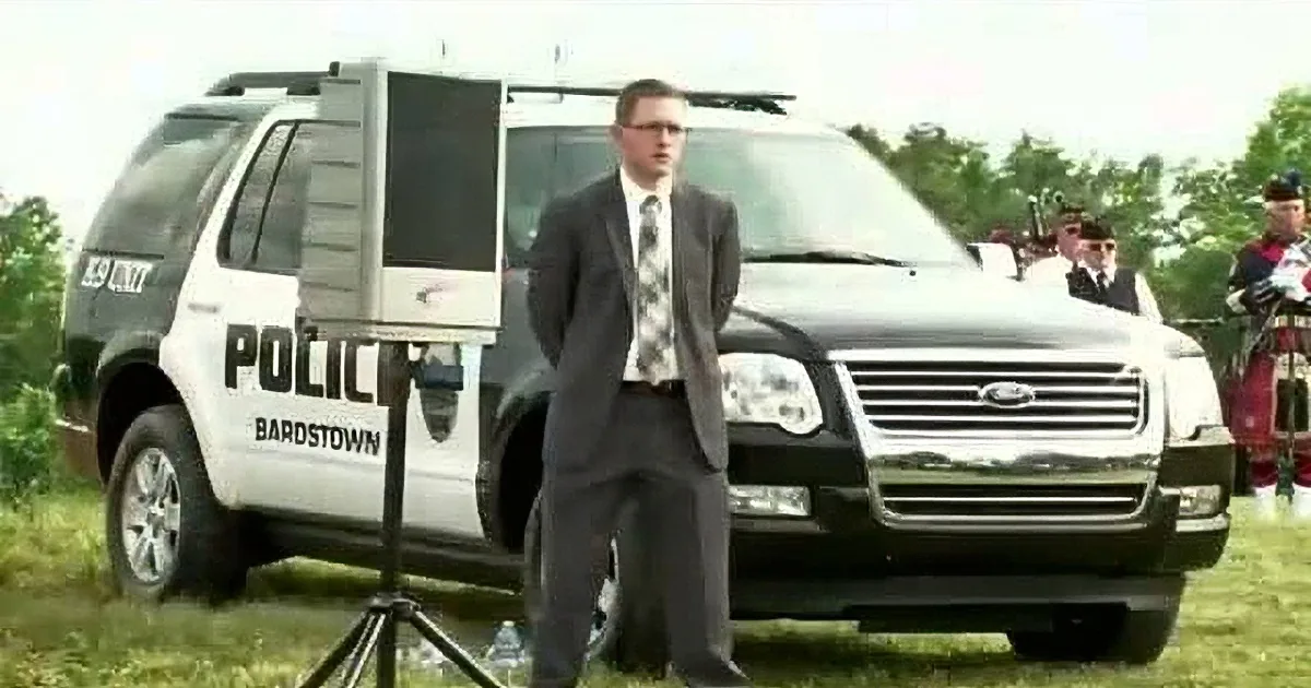 A man in a suit stands in front of a police SUV, with another vehicle and people in the background.