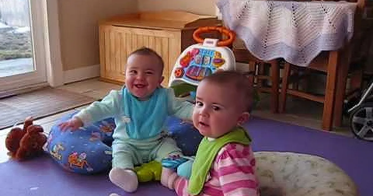 Two smiling babies sit on a purple mat with toys and furniture around them.