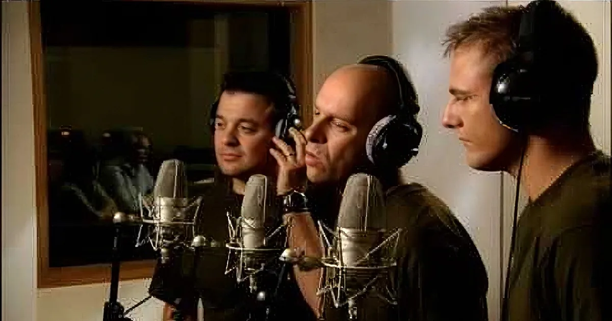 Three men in a recording studio, wearing headphones, focused on microphones.