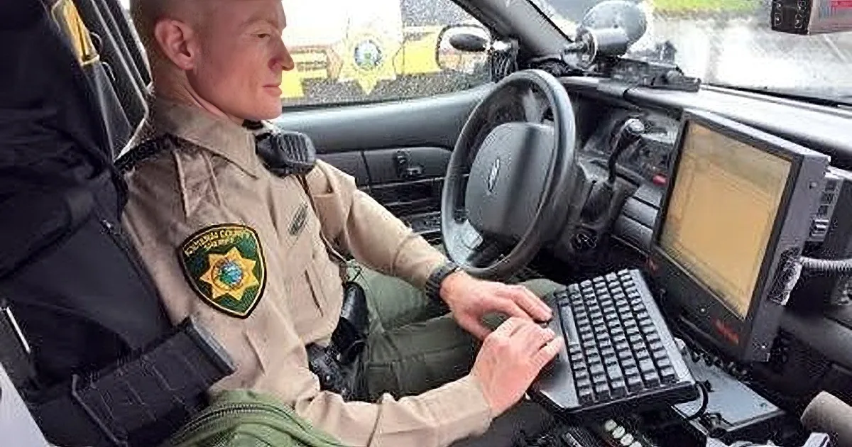A police officer in uniform sits at the steering wheel of a patrol car, holding a device.