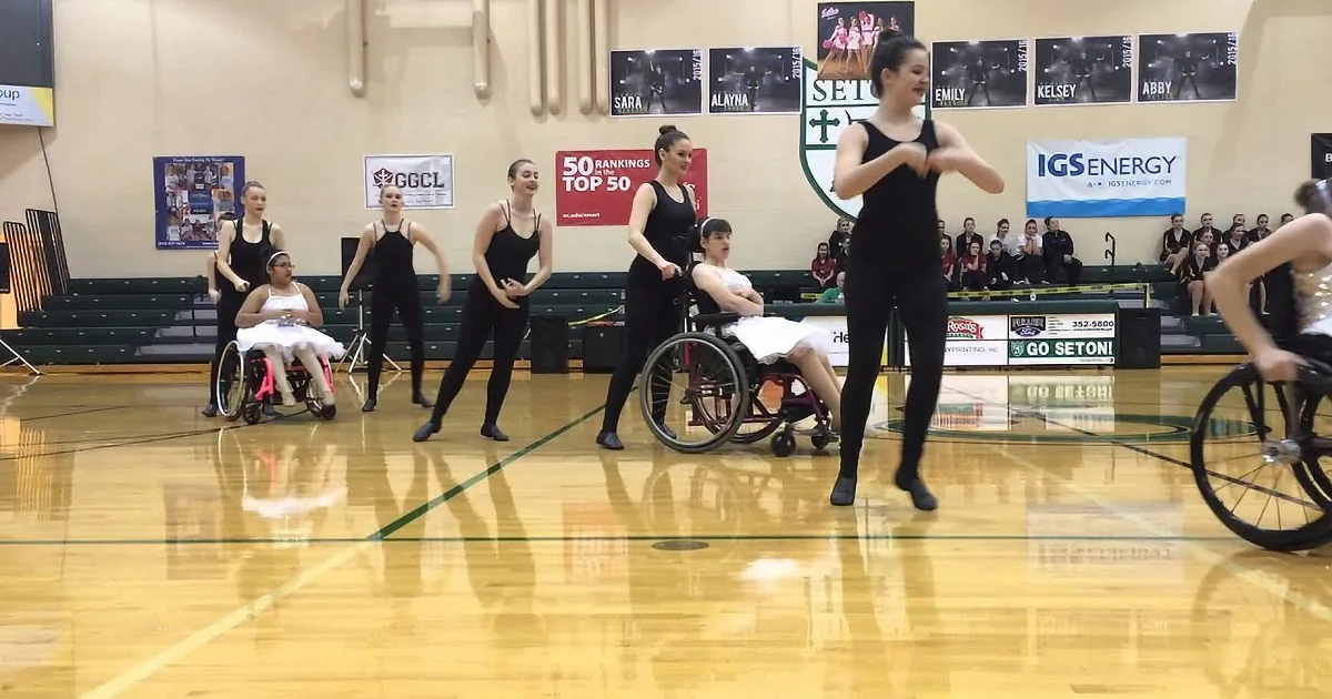 A dancer in a wheelchair performs with others in a gymnasium, surrounded by spectators and banners.