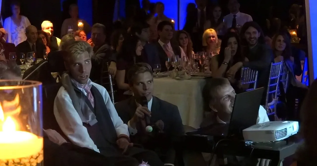 A man in a suit sits at a formal event, surrounded by a crowd, with a projector and screen in the foreground.