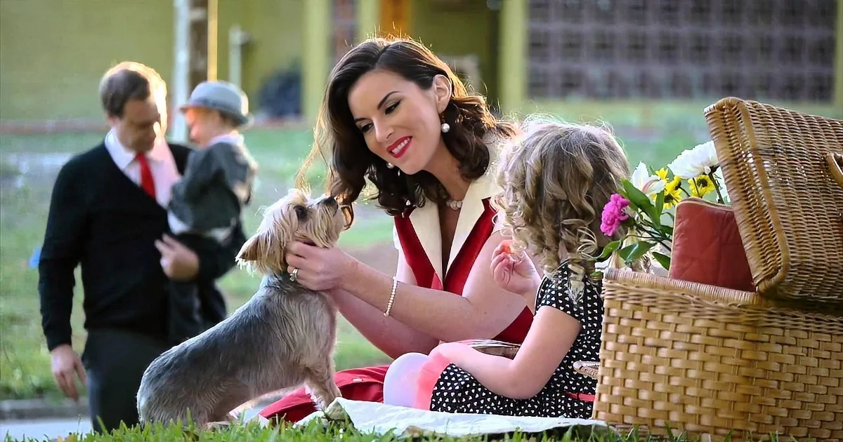 A woman in a red dress pets a small dog at a picnic, surrounded by greenery and picnic baskets.