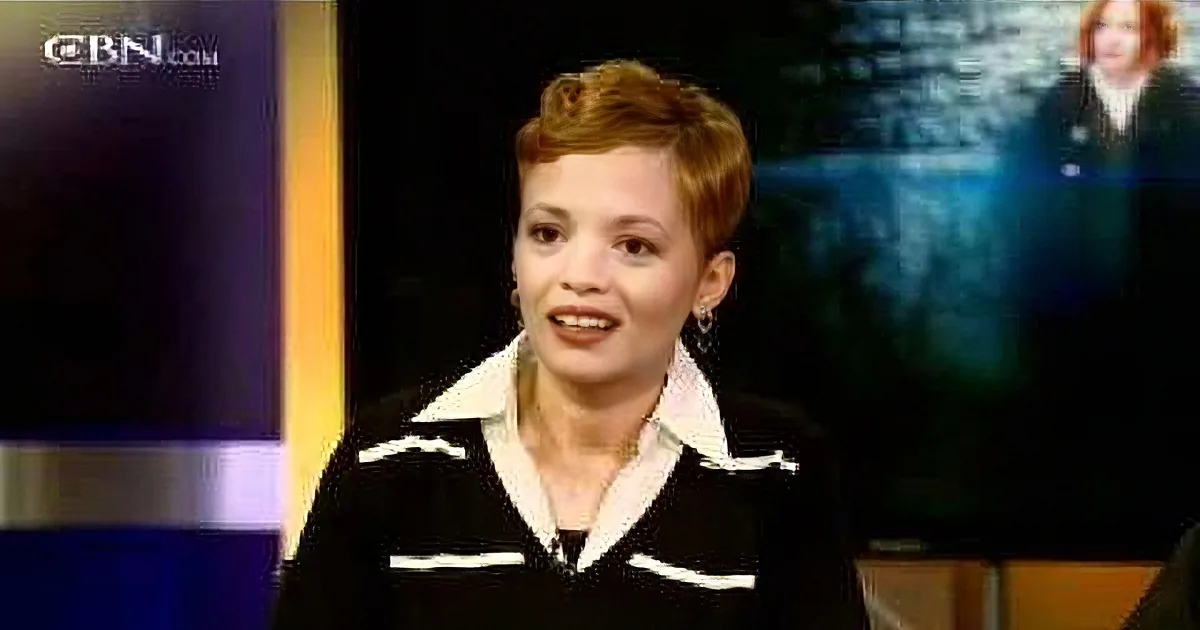 A woman with short hair, wearing a black and white outfit, smiles while speaking on a television show.
