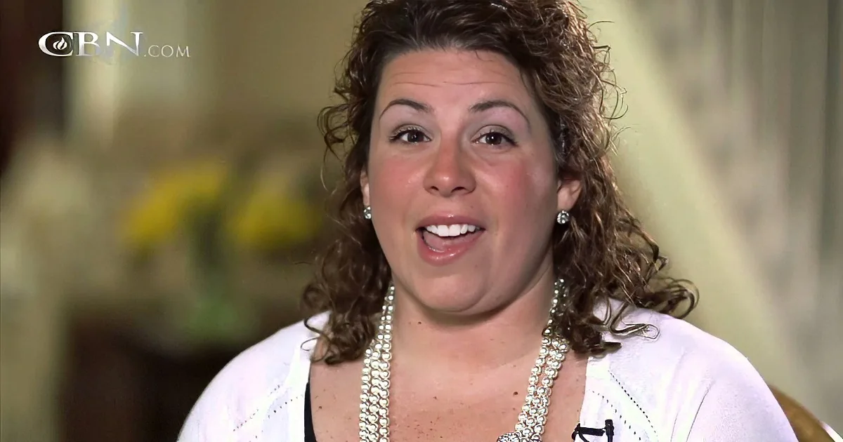 A woman with curly hair, wearing a white cardigan and pearl necklace, appears to be speaking.