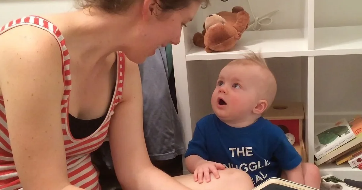 A woman in a red and white striped tank top reads to a baby in a blue shirt, both seated on the floor.