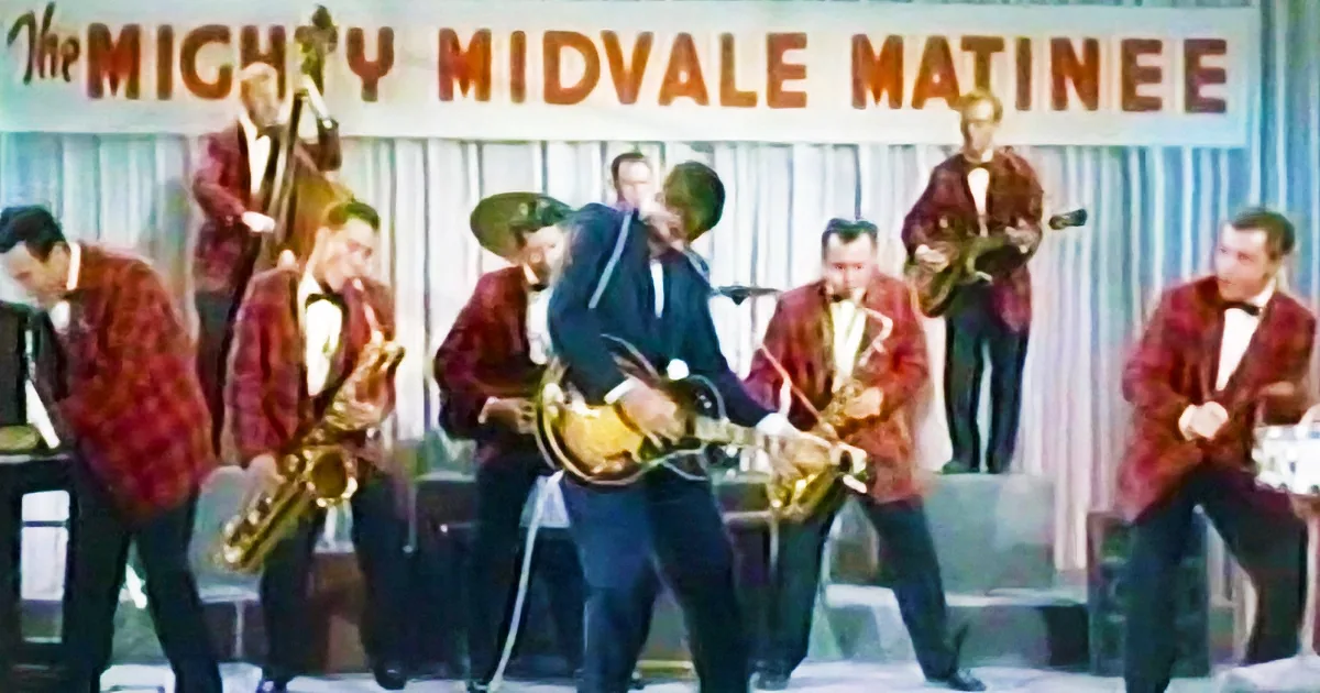 A guitarist in a blue suit plays a golden guitar on stage with a band in red plaid jackets.