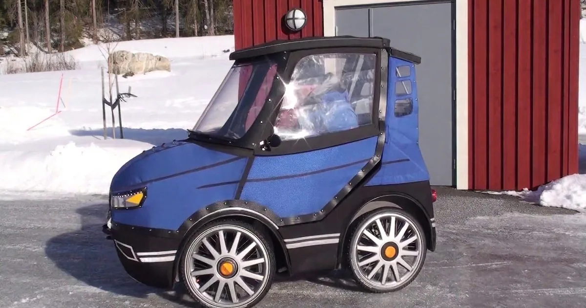 A small, blue, futuristic-looking vehicle with large wheels is parked in front of a red building on a snowy day.