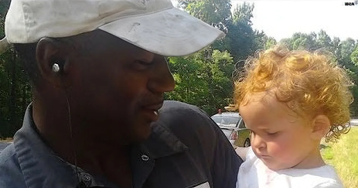 A man wearing a cap and earphones interacts with a child with curly hair, both outdoors.