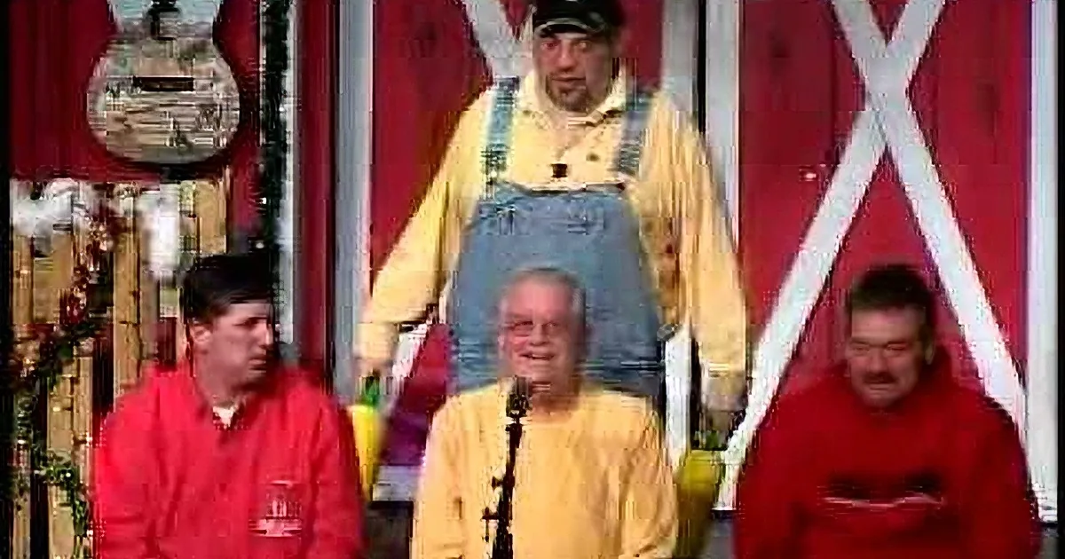 A man in overalls and a yellow shirt stands on stage, surrounded by others in red and yellow attire, with a barn backdrop.
