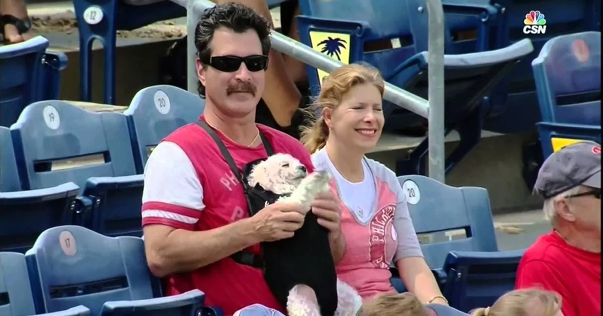 A man holds a small white dog while sitting in a stadium with a woman beside him.