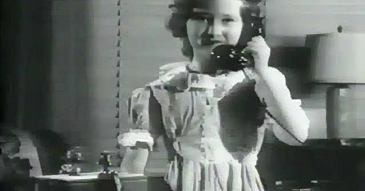 A woman in a dress is holding a phone to her ear, standing in a kitchen with pots and a window in the background.