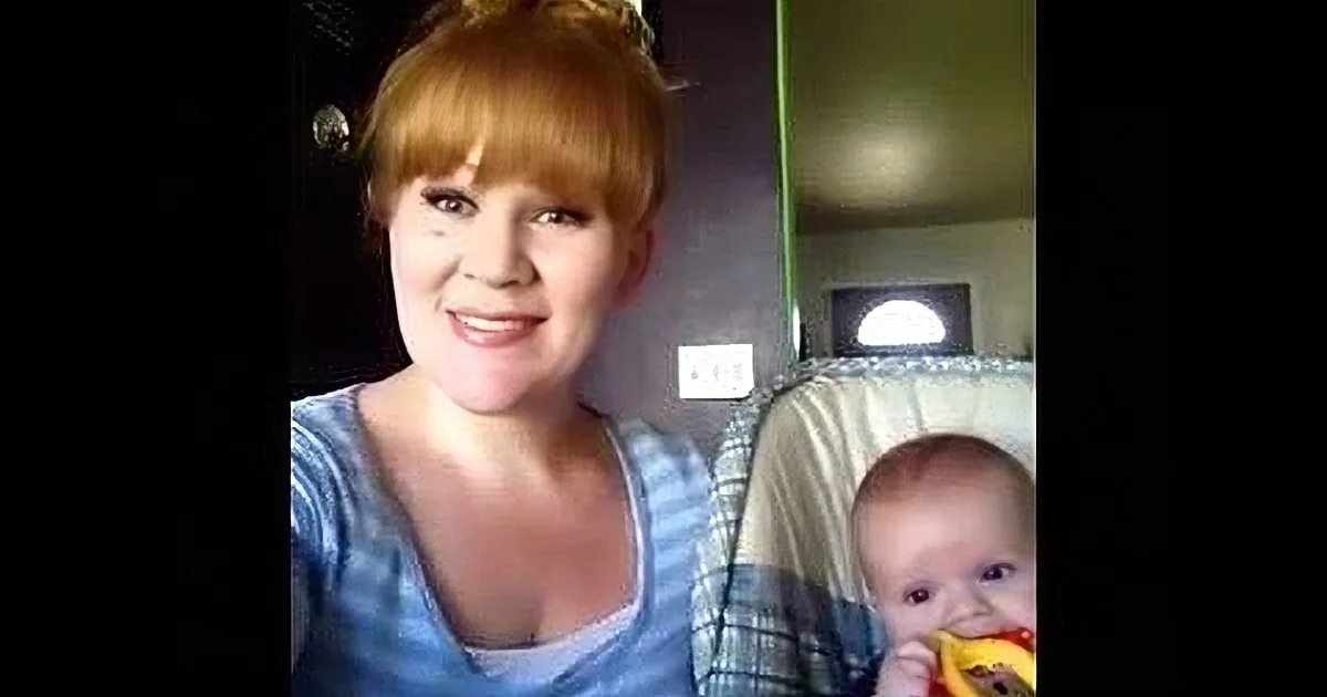 A woman with red hair and a blue striped shirt is smiling, with a baby and another person in the background.