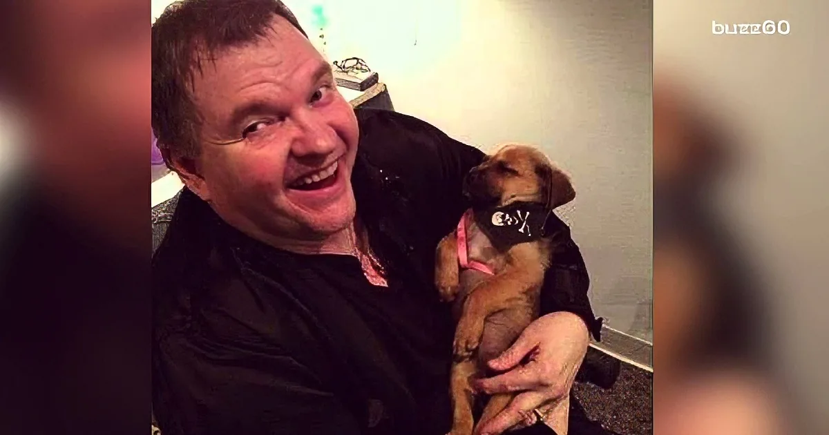 A man in a black shirt is holding a small brown puppy with a pink collar, both smiling.