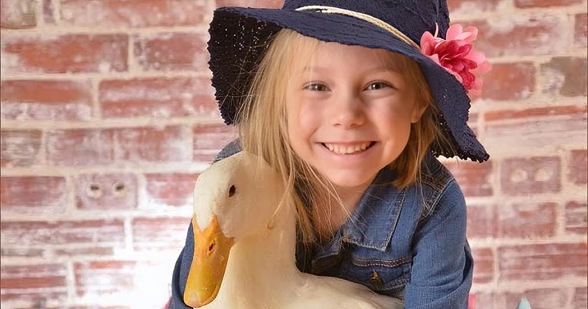 A smiling girl in a denim jacket and hat holds a duck against a brick wall backdrop.