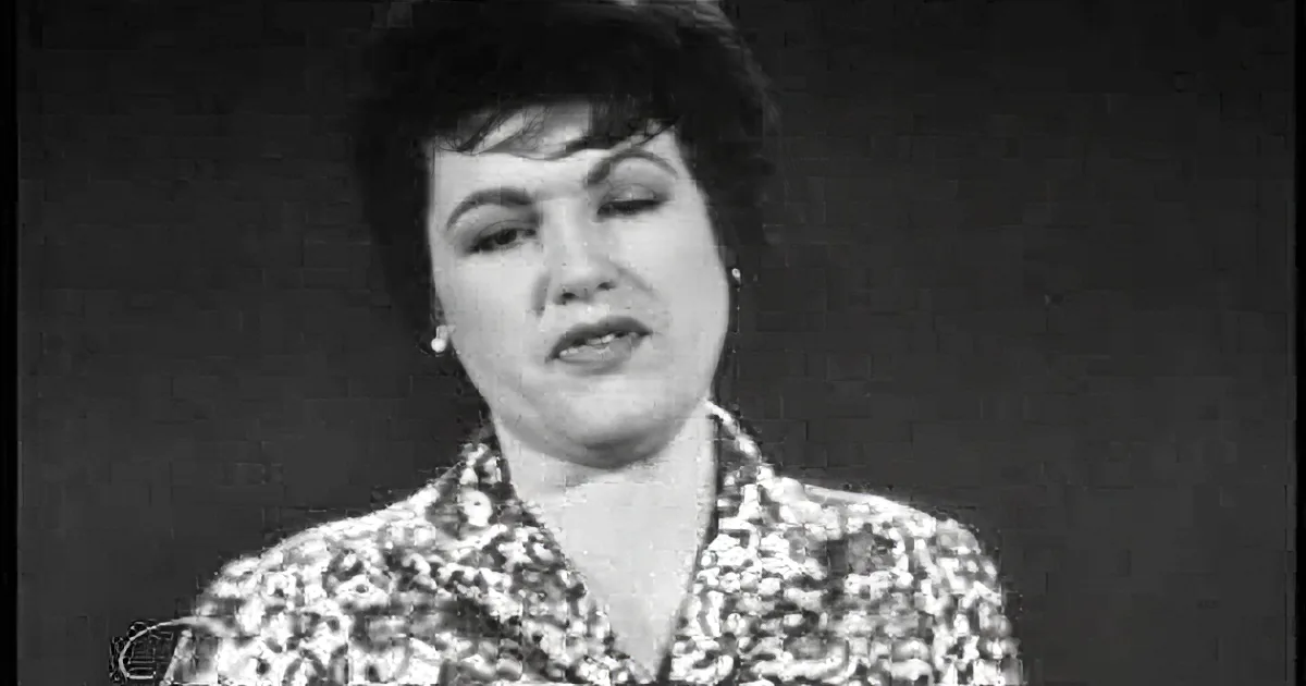 A woman in a patterned blouse sings passionately with her eyes closed, captured in a black-and-white image.