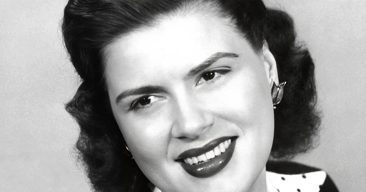 A woman with dark, curly hair and a polka-dotted blouse smiles warmly.