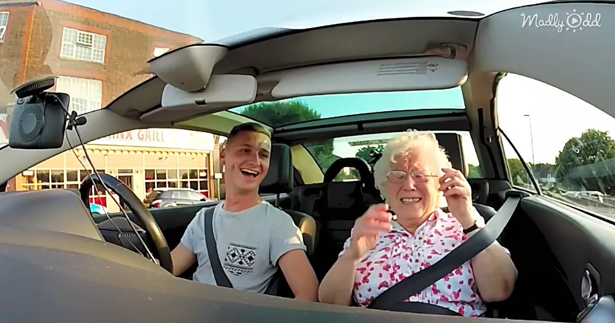 A young man and elderly woman are smiling in a car, both wearing seatbelts, with a building and trees visible outside.