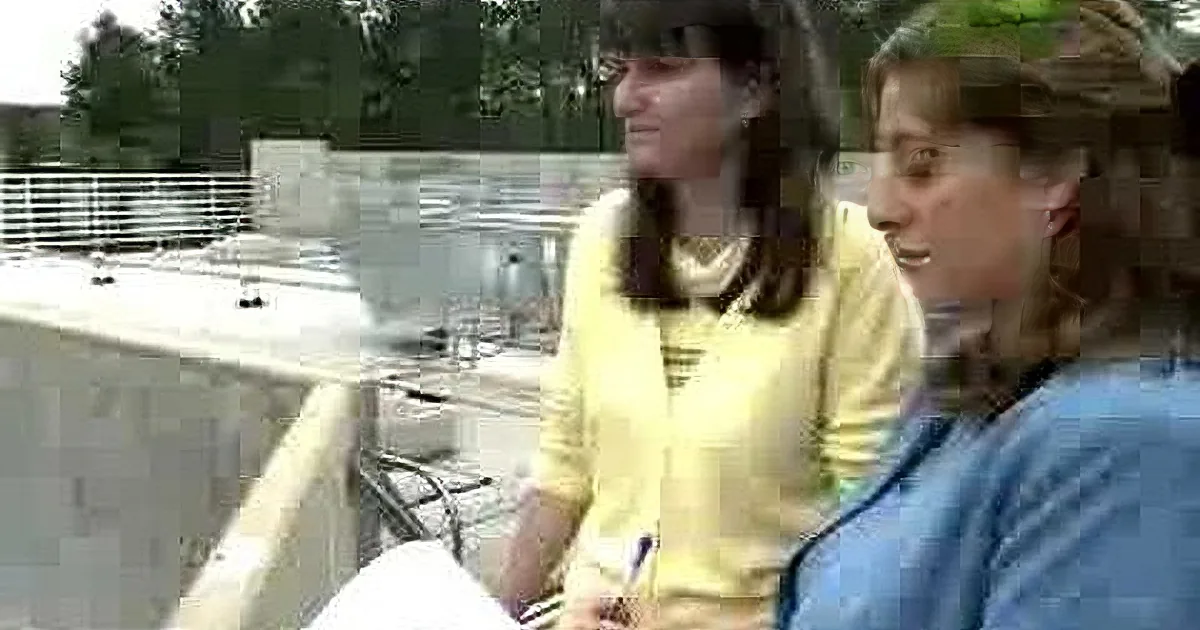 Two women are sitting outdoors, engaged in conversation, with a concrete structure and railing in the background.