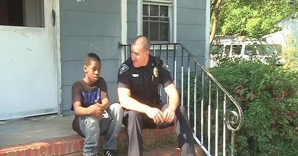 A police officer and a boy sit on a step outside a house, engaging in conversation.