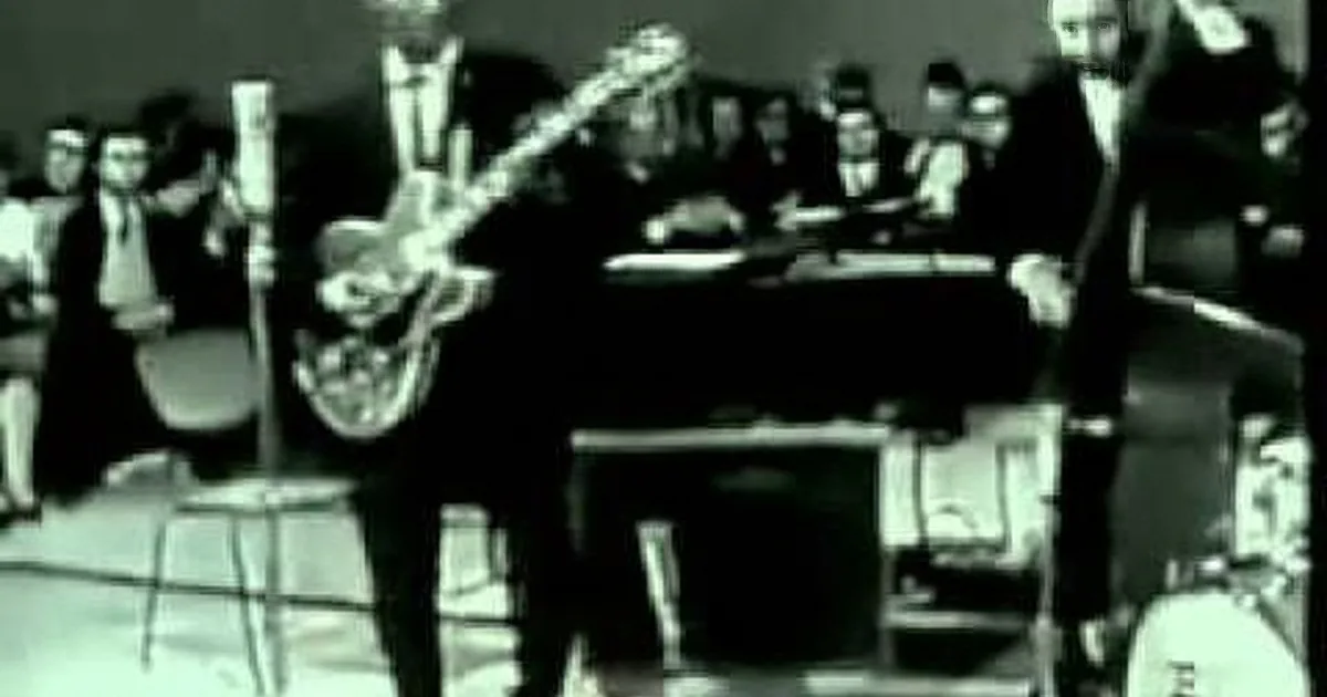 A guitarist in a suit plays an electric guitar on stage, surrounded by an audience and other musicians.