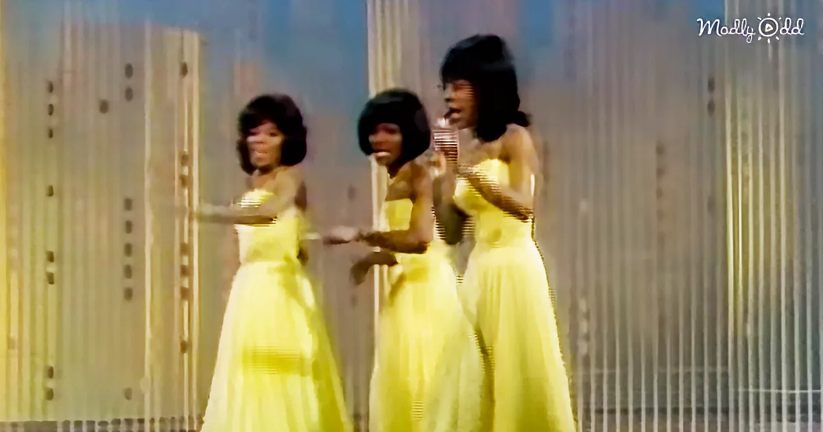 Three women in yellow dresses perform on stage with a cityscape backdrop.