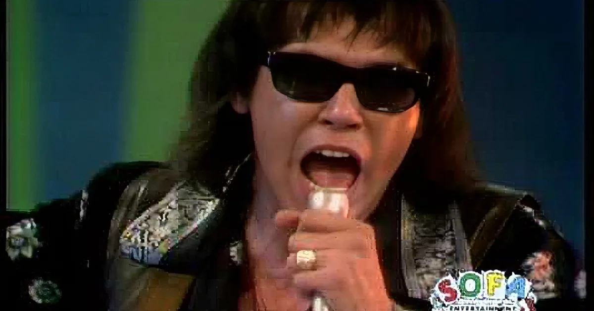 A person in a black leather jacket and sunglasses is singing passionately into a microphone.