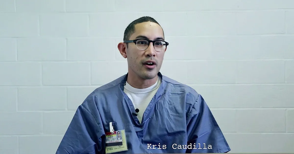 A man in scrubs with a name tag, Kris Caudilla, is speaking in front of a white brick wall.