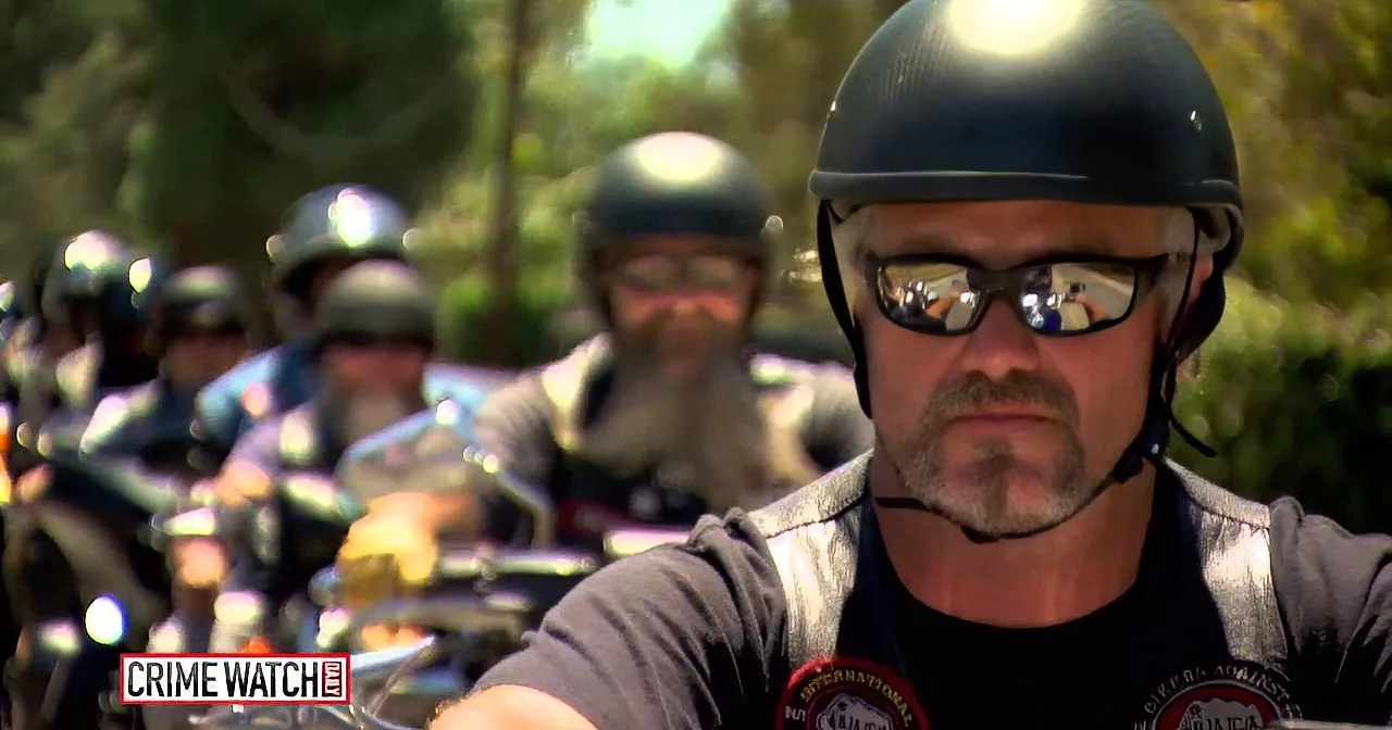 A man in a black helmet and sunglasses rides a motorcycle, leading a group of similarly dressed riders.