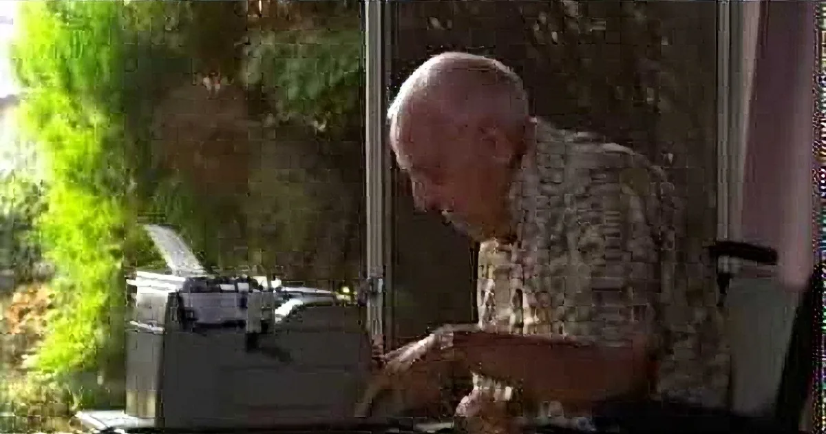 An older man with a bald head and glasses is sitting at a desk, typing on a typewriter.