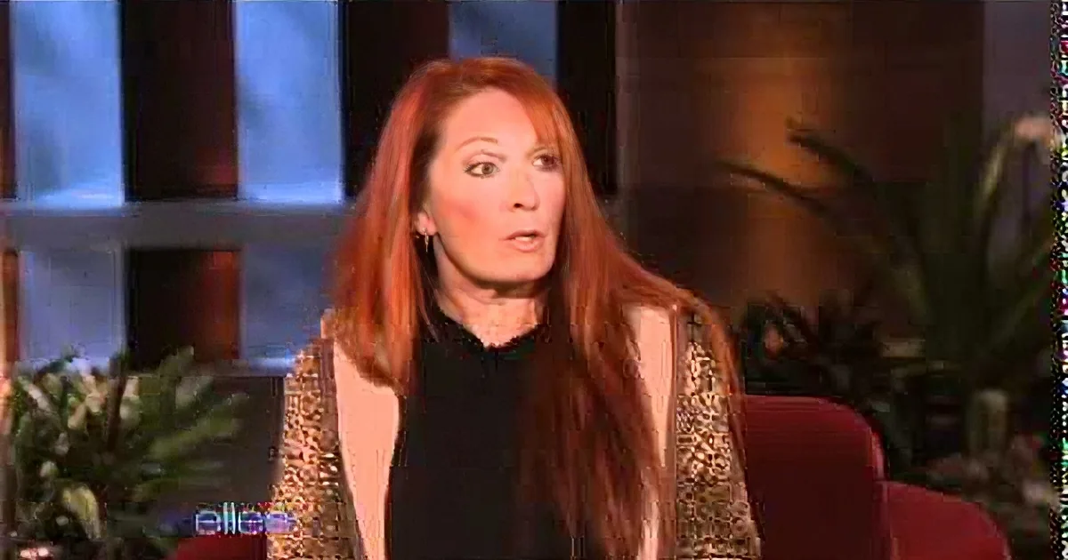 A woman with long red hair, wearing a black and gold outfit, sits in a studio setting with a blurred background.