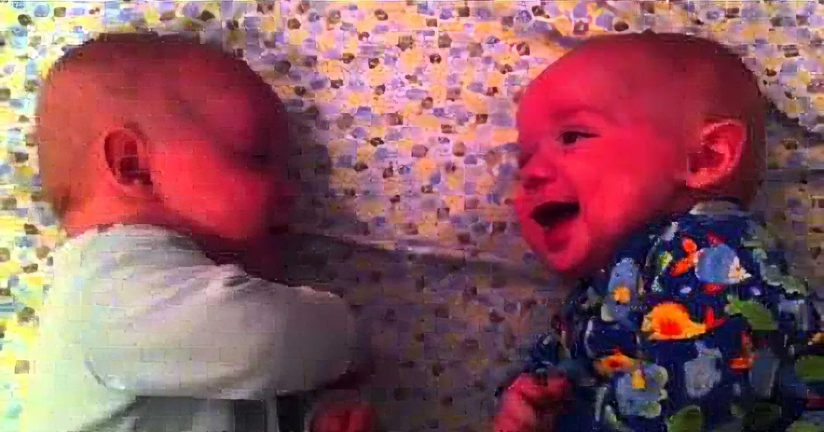 Two babies are playfully interacting, one with a spoon in its mouth, both appearing happy and engaged.
