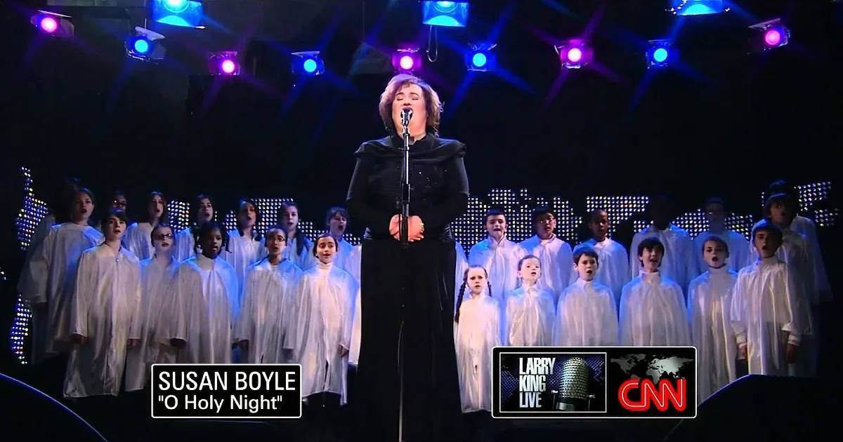 Susan Boyle sings "O Holy Night" on stage with a choir, under colorful stage lights.